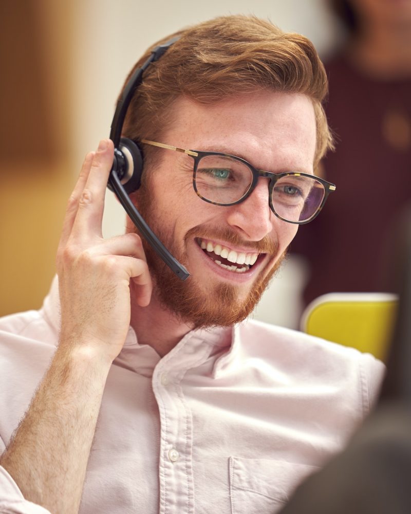 Businessman Wearing Headset Talking To Caller In Busy Customer Services Centre